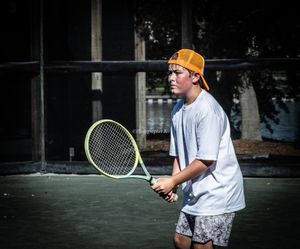 Tennis player in white shirt and orange cap practices backhand swing technique on outdoor court.