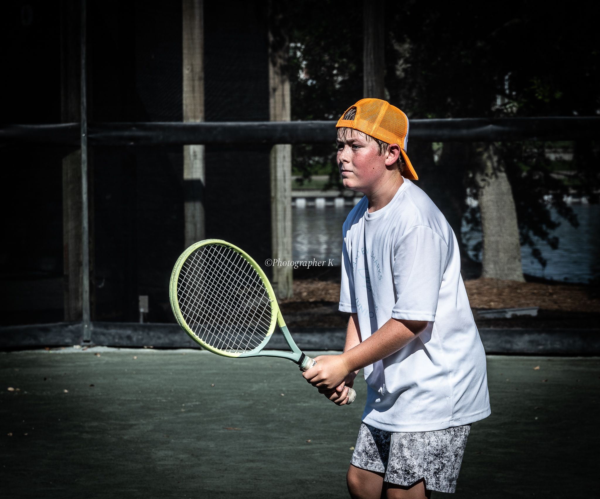 Tennis player in white shirt and orange cap practices backhand swing technique on outdoor court.