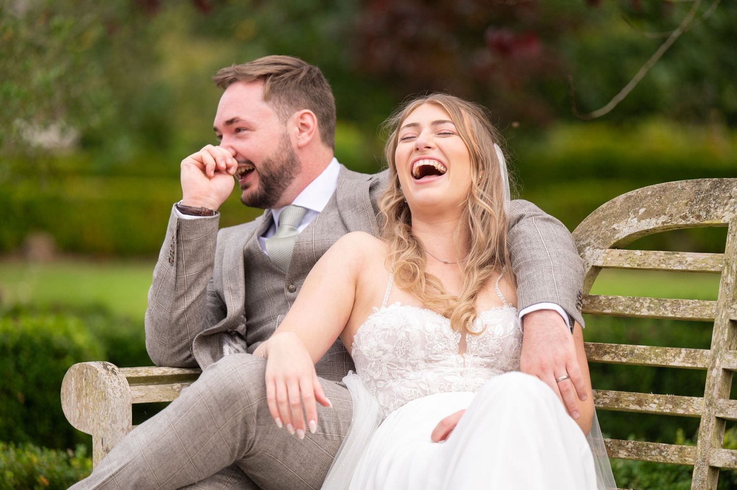 A bride and groom share a candid laughing moment together while sitting on a wooden bench in a garden setting.
