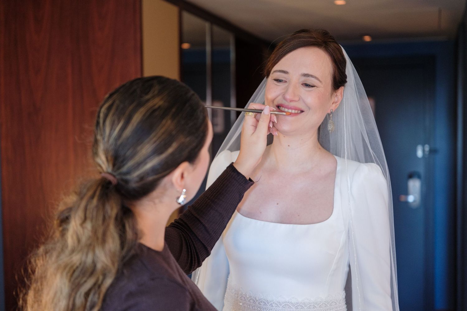 A bride in a white sleeveless wedding dress and veil smiles during her wedding preparations.