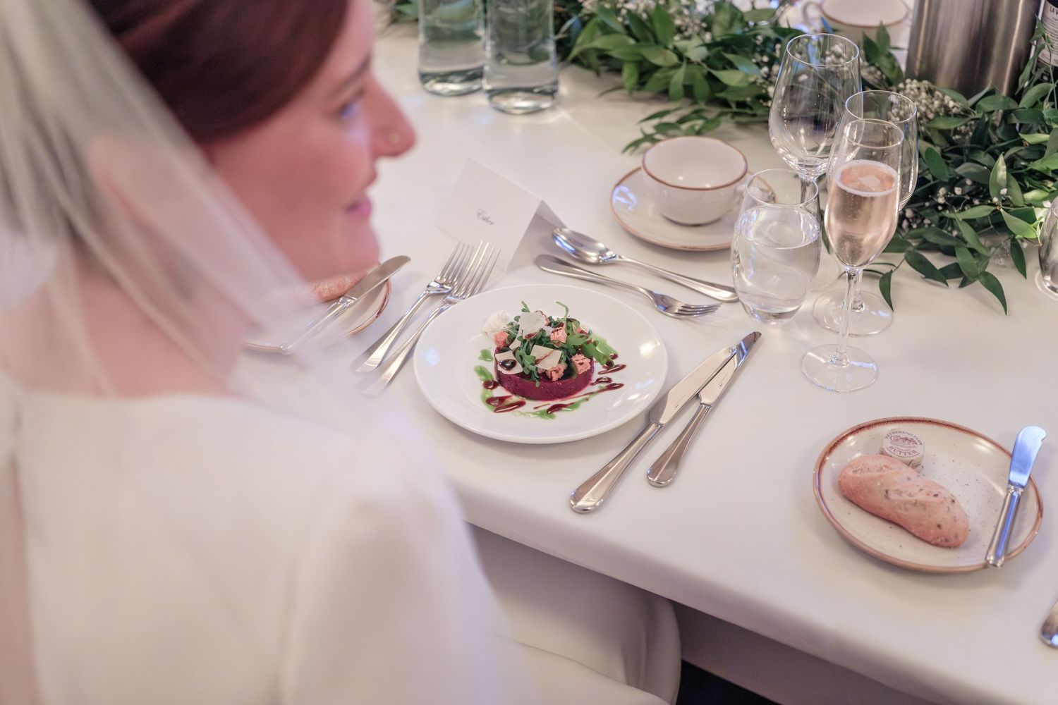 An elegant wedding reception table setting with a plated salad, bread, and greenery garland decorations.