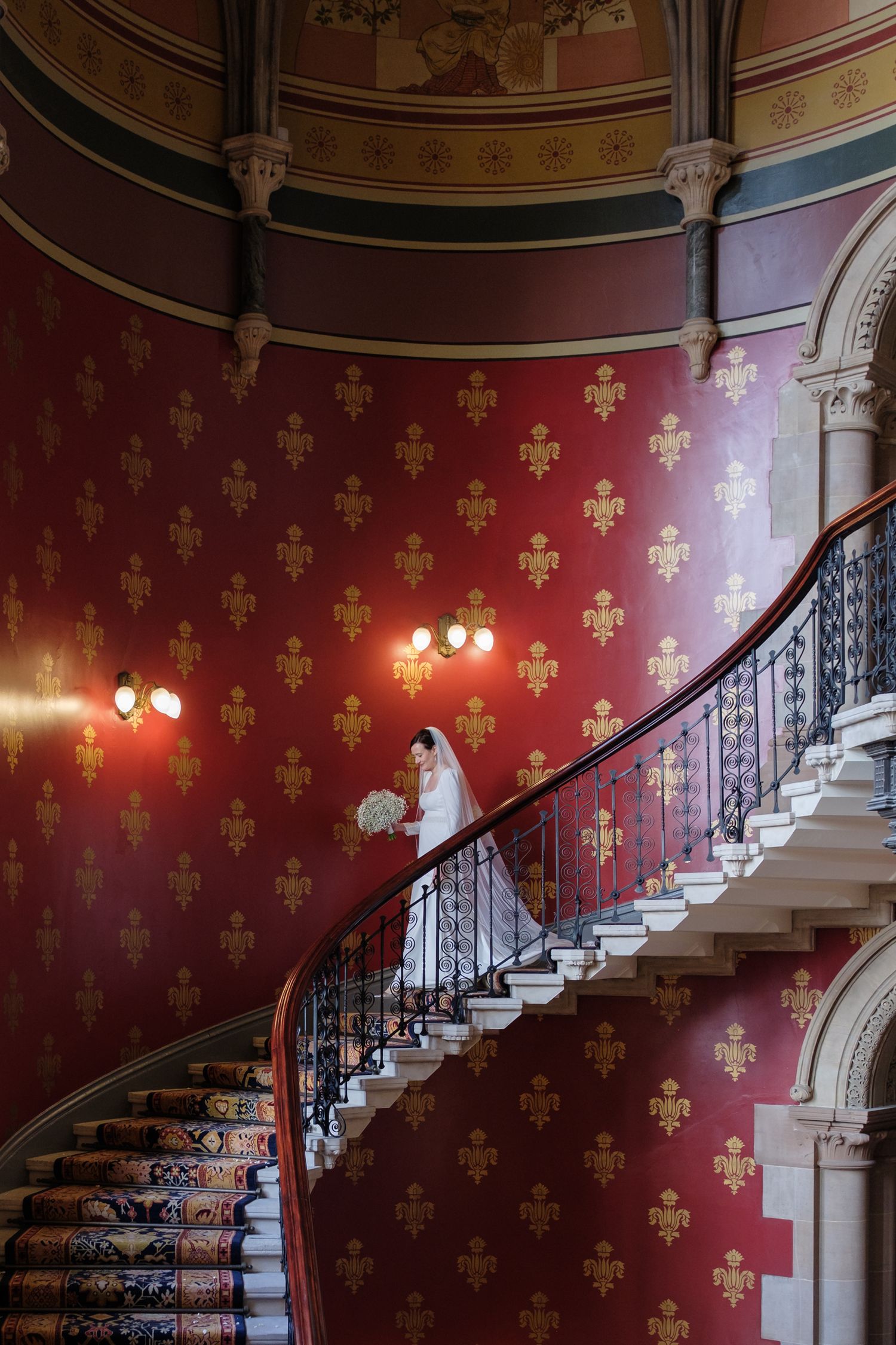 Elegant curved staircase with ornate iron railing against deep red wallpaper featuring fleur-de-lis pattern in historic interior.