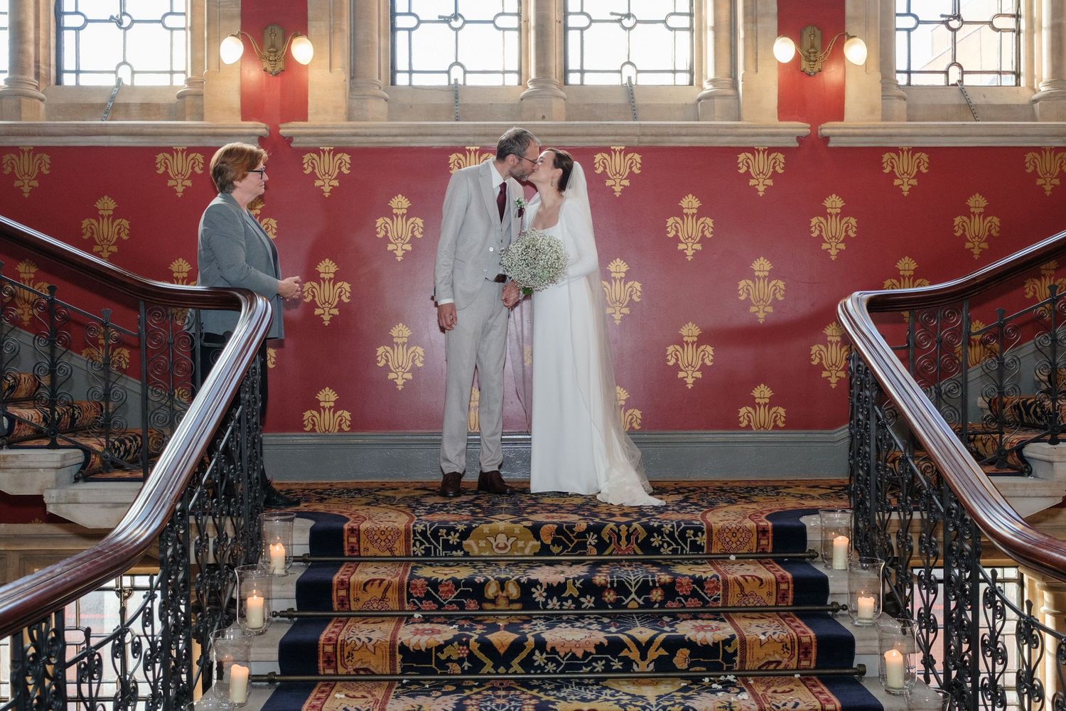 A couple stands on ornate red carpeted stairs in a historic building during their wedding ceremony.