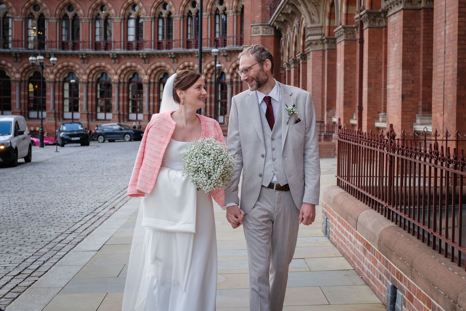 A couple walks together in formal wedding attire along a historic brick building with arched windows and iron railings.