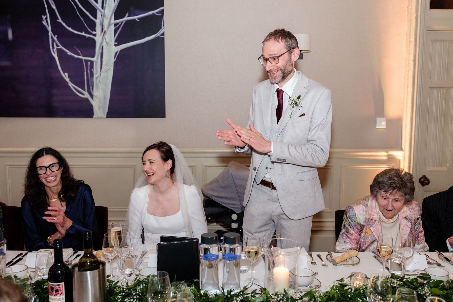 Wedding reception scene with guests seated at a decorated table while someone gives a speech.