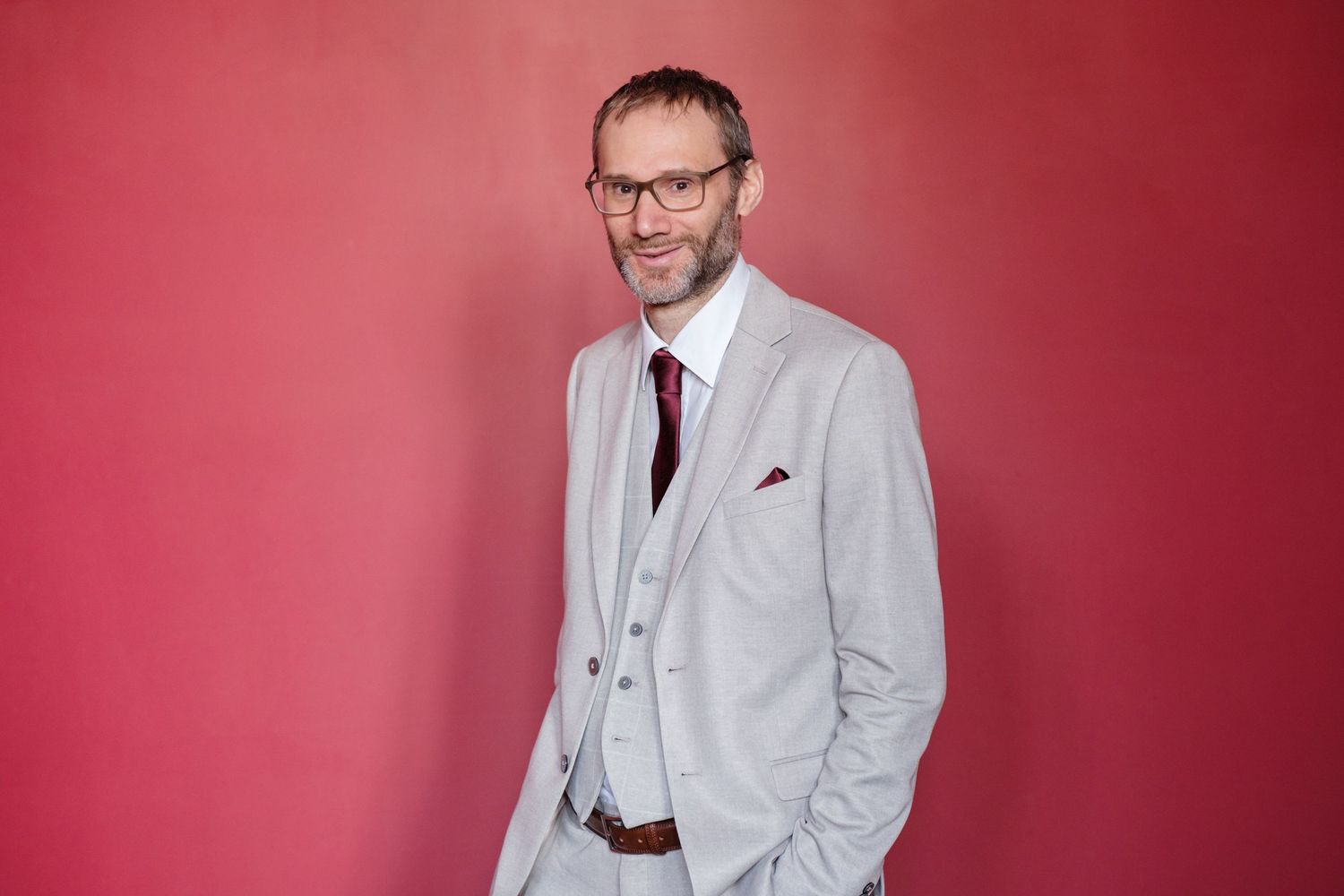 A person wearing a light gray suit and glasses poses confidently against a red background.