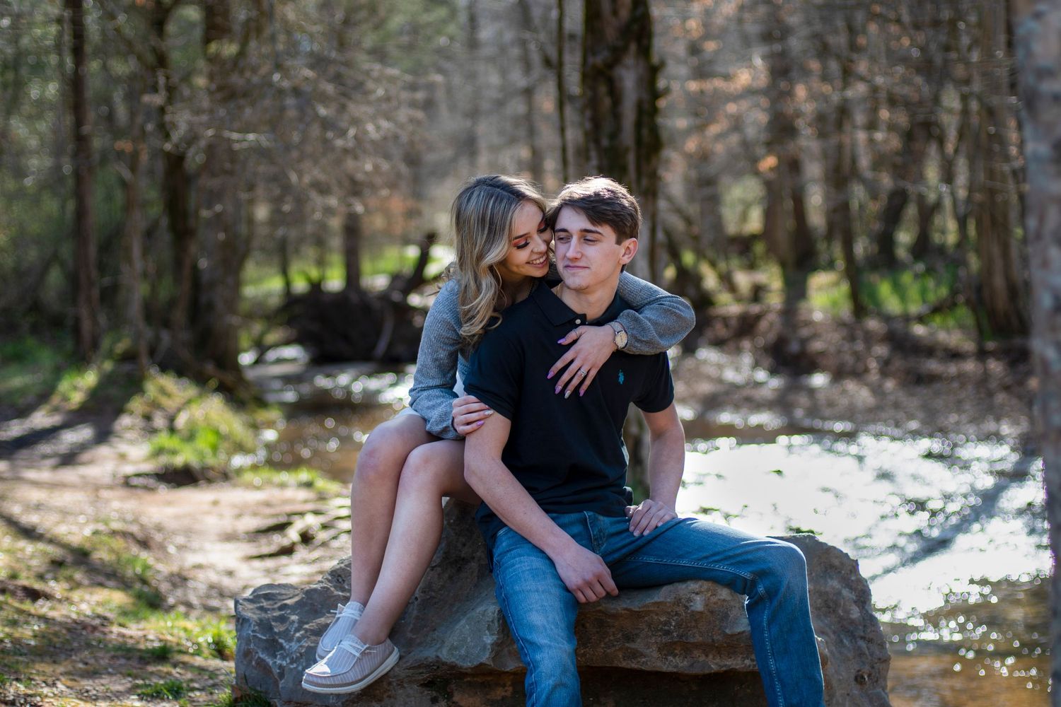 A couple sits together on a rock by a creek in a wooded setting.