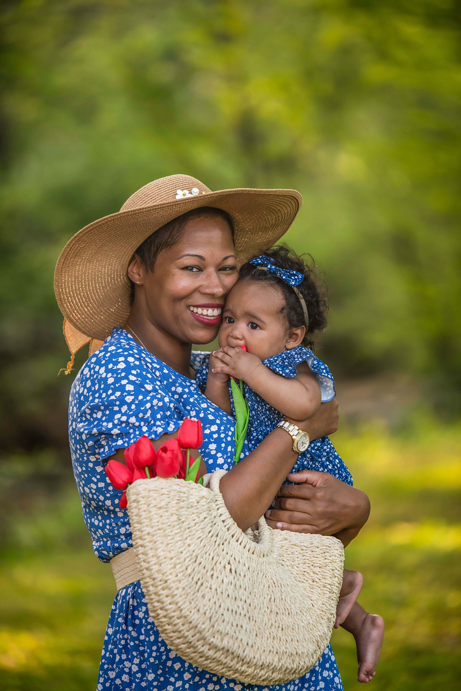Woman in blue floral dress and sun hat holds baby while carrying straw bag with tulips.