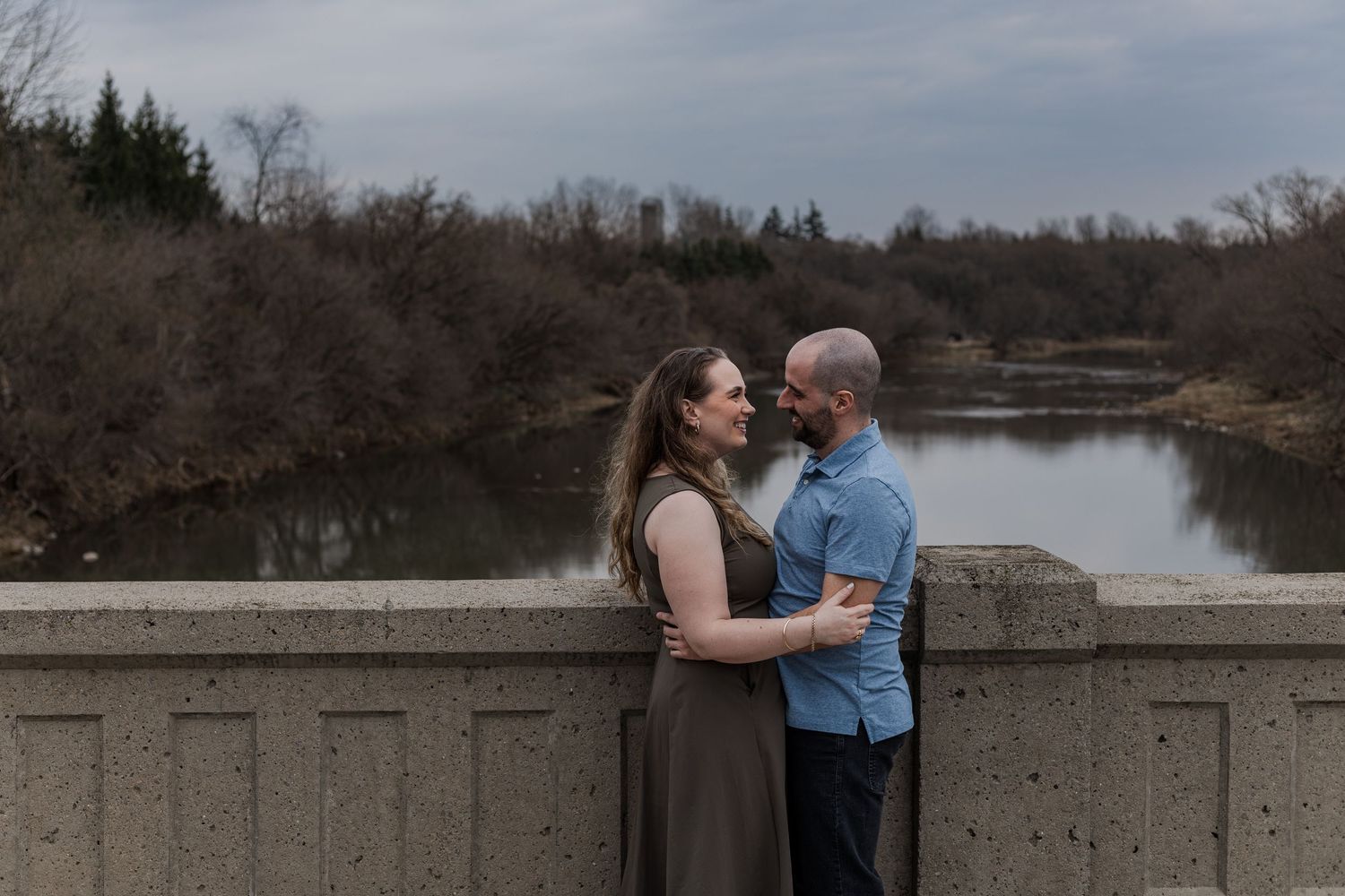 A couple shares a romantic moment together on a bridge overlooking a serene lake on an overcast day.