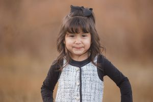 A child wearing a black and white dress poses outdoors in autumn with a blurred brown background.