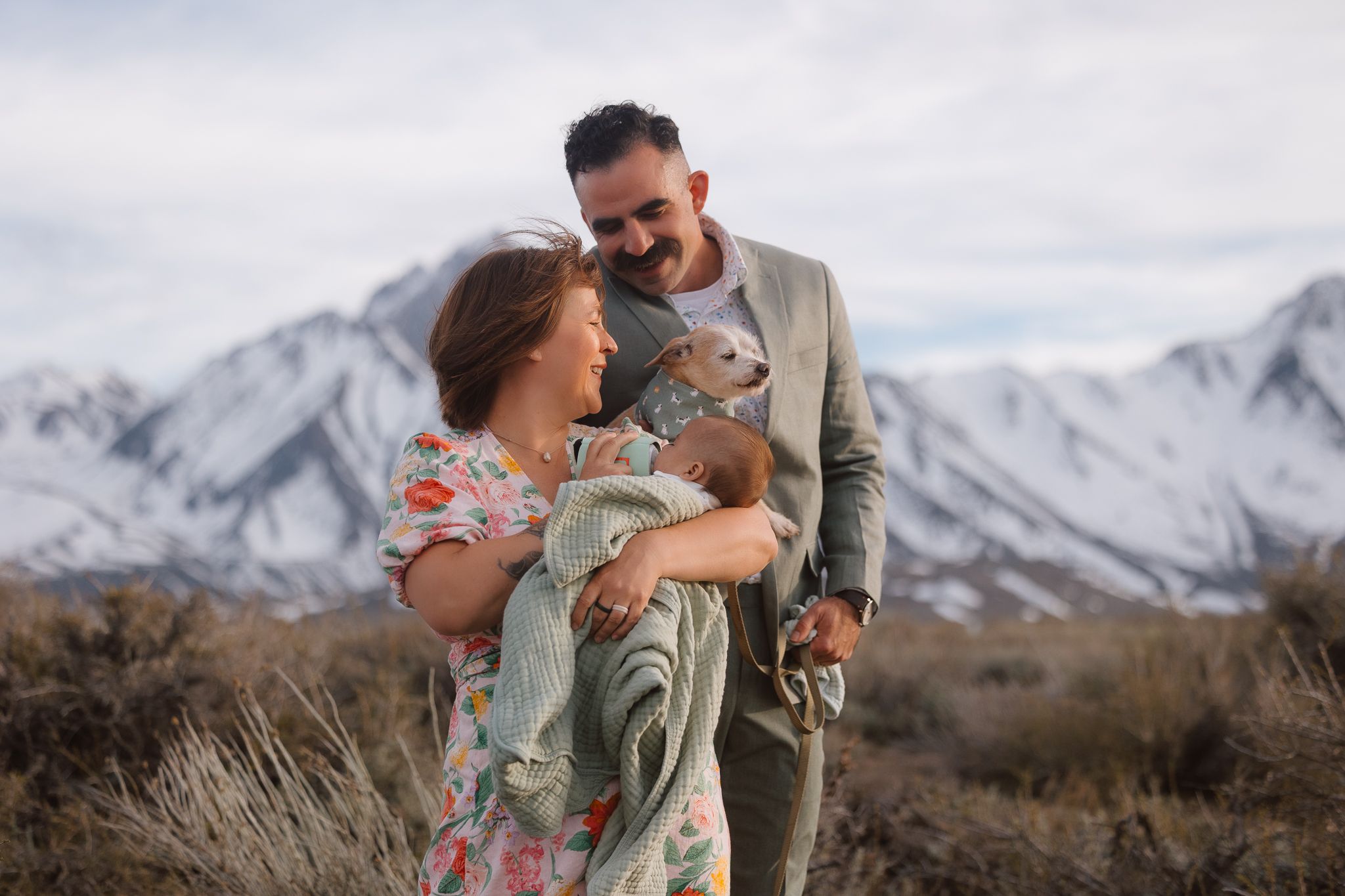 A happy couple holding their newborn baby outdoors with snow-capped mountains in the background.