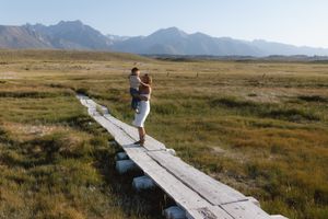 Woman holding a child standing on a wooden boardwalk path through green meadows with mountain range backdrop.