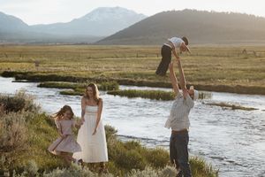 Family enjoying outdoor time by a mountain stream, with father tossing child in air while mother and daughter watch.