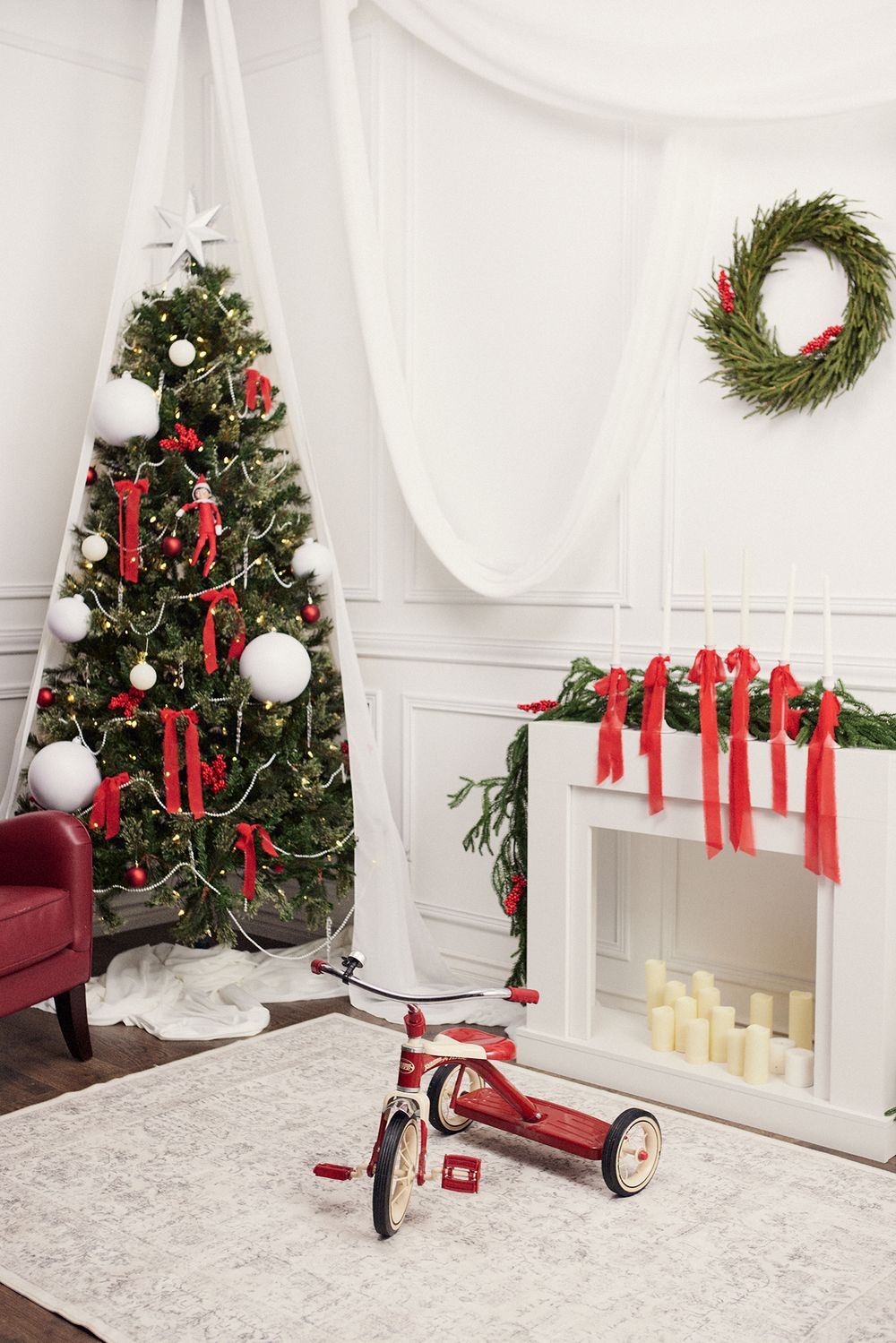 White Christmas interior with decorated tree, wreath, and red tricycle by fireplace.