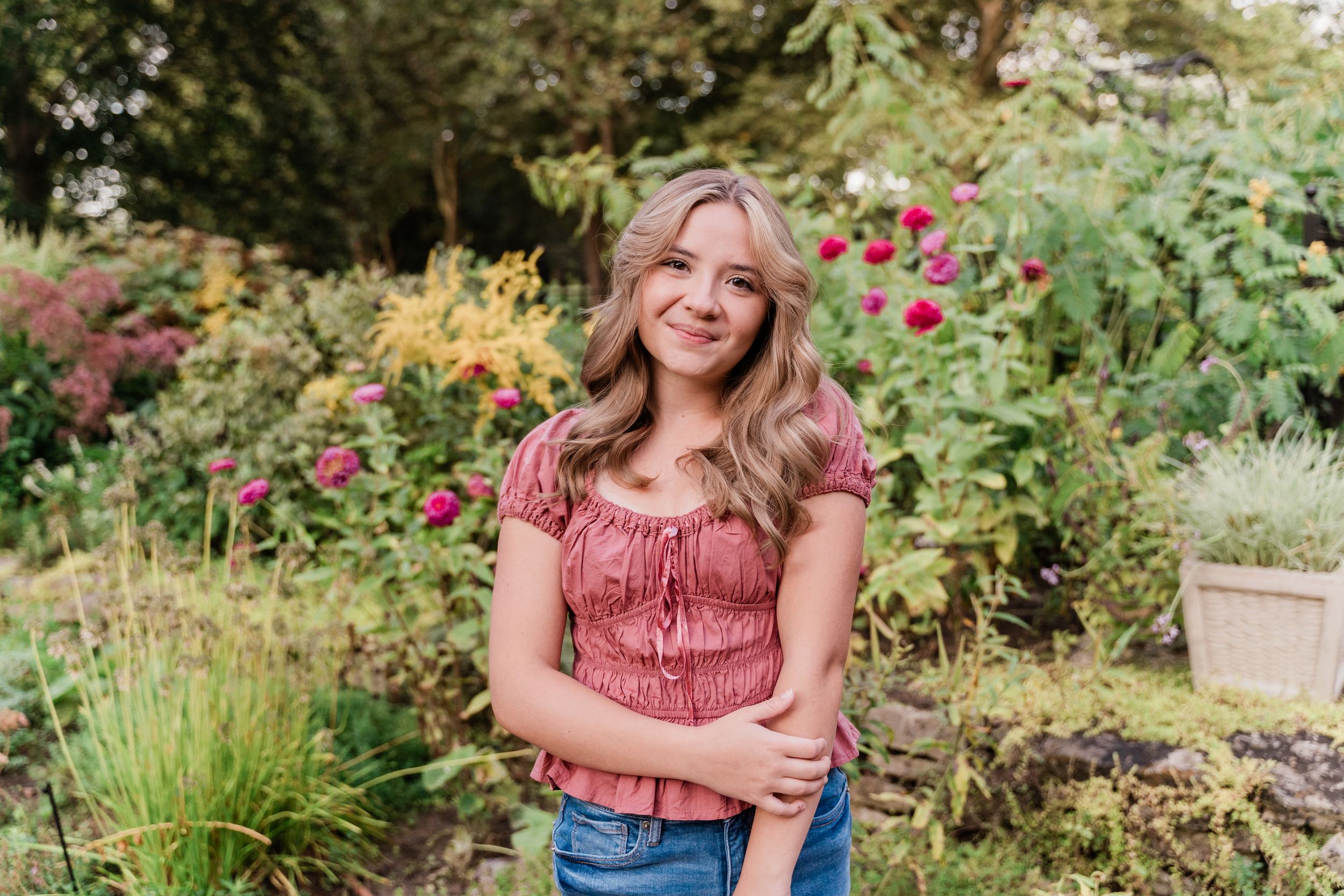 A person in a red top and jeans sits in a lush garden surrounded by blooming flowers and greenery.