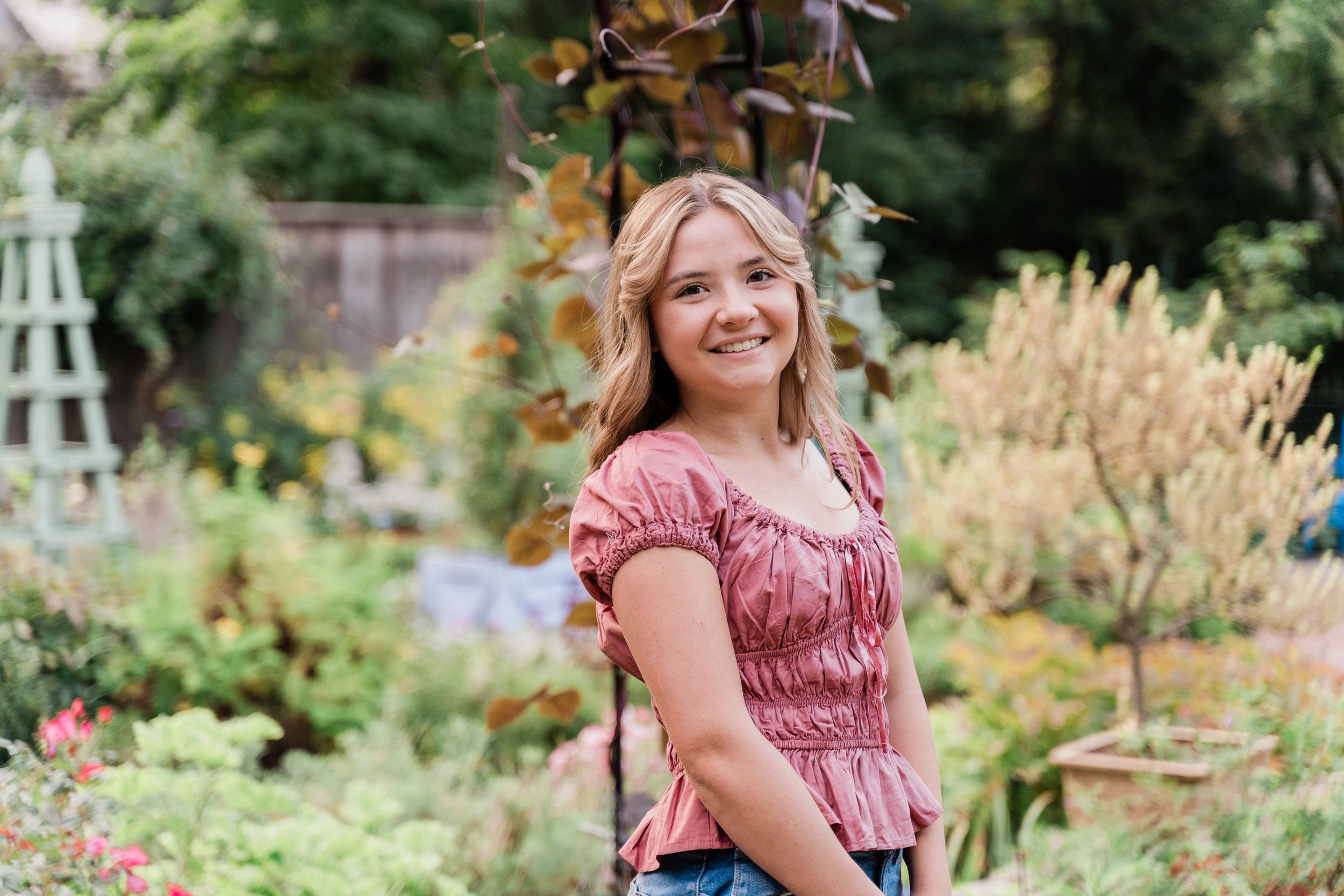 A person in a pink ruched top sits in a lush garden setting surrounded by summer flowers and greenery.