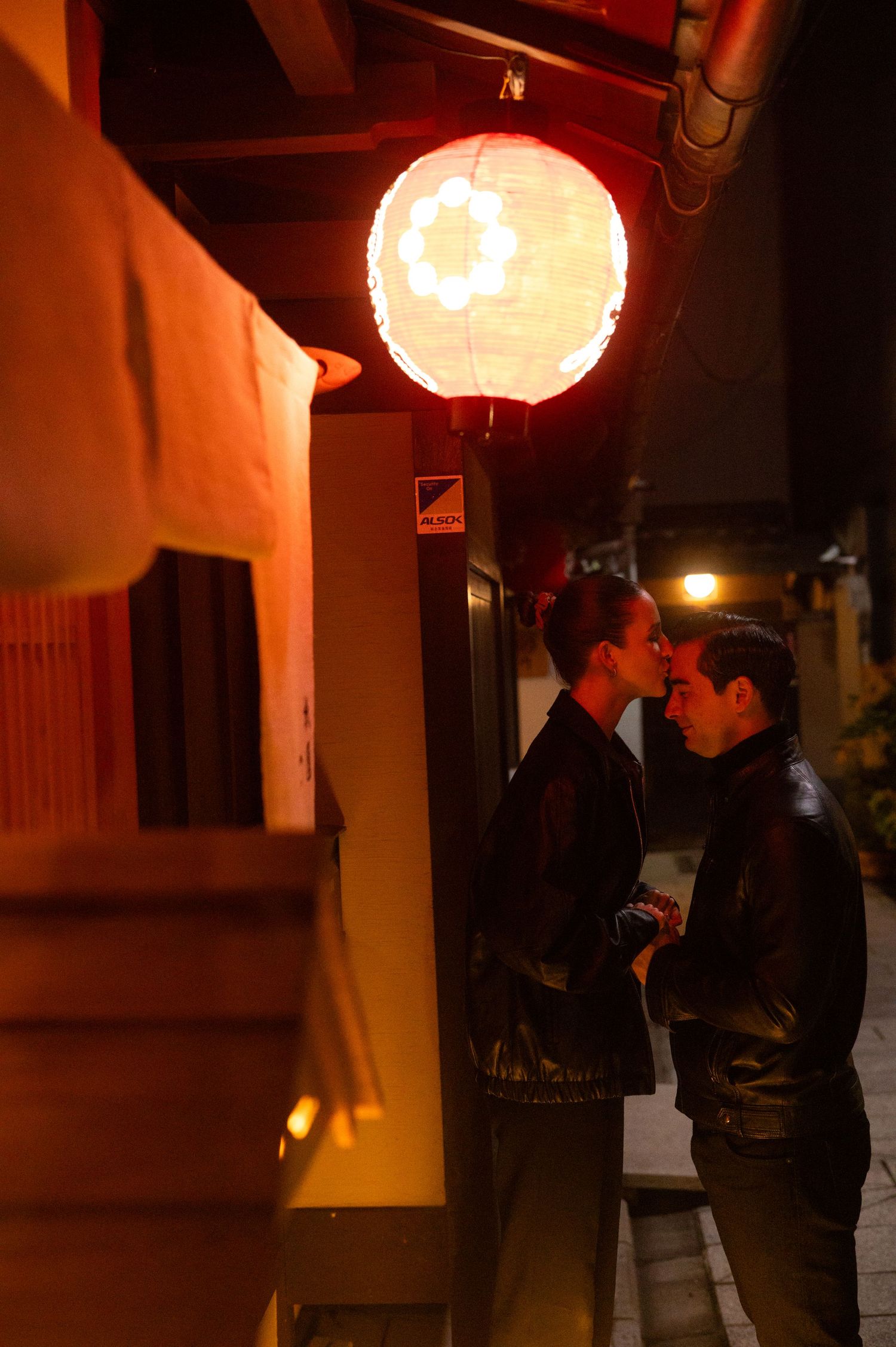Couple almost kissing beneath a glowing paper lantern on a Japanese street at night.