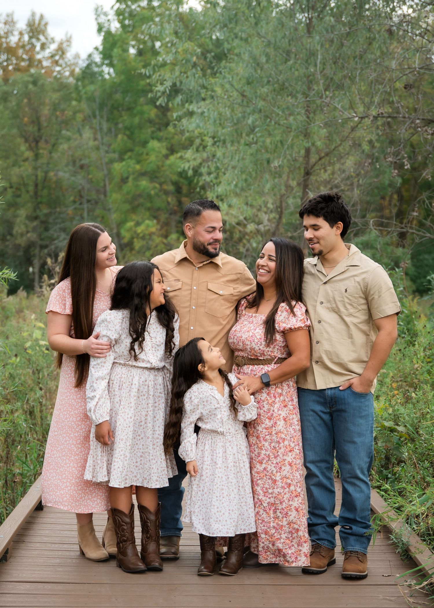 A family laughs together while standing on a wooden boardwalk surrounded by lush greenery in matching casual outfits.