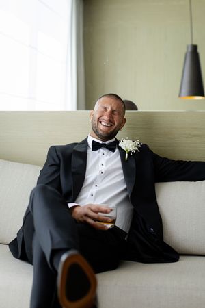 A person in a black tuxedo lounges on a light-colored couch while laughing and relaxing during a formal event.