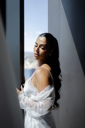 A bride in a delicate white lace gown and robe poses near a window with dramatic natural lighting.