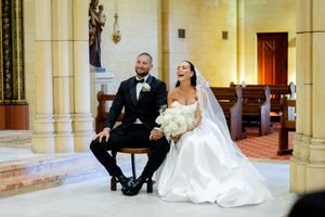 A joyful wedding couple sits and laughs together on elegant stone steps in a grand hallway.