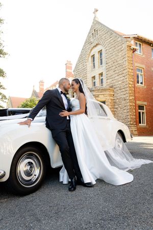 Romantic wedding couple poses with classic white vintage Rolls Royce car outside stone building.