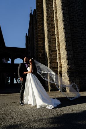 Bride and groom pose for dramatic wedding photos under stone archways at sunset, with veil flowing in the wind.