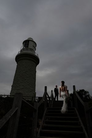 A lighthouse silhouetted against a moody gray sky with figures ascending stone steps in the foreground.