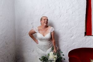 A joyful bride in a white lace dress laughs while holding a bouquet against a white wall with red accents.
