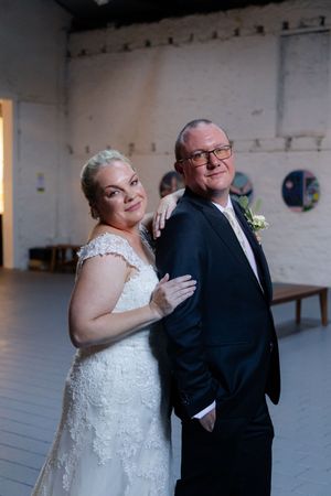 A bride in a white lace dress shares a sweet candid moment with someone in a dark suit at an indoor venue.