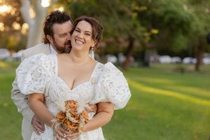 A couple in white wedding attire share a joyful moment together outdoors while holding an autumn-colored bouquet.