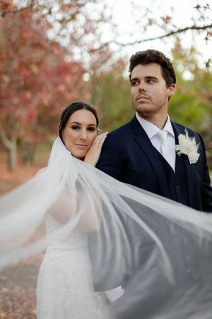 A bride and groom pose outdoors in autumn with her flowing white veil catching the breeze among fall foliage.