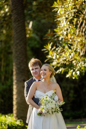 A joyful wedding couple shares a candid moment together in a sun-dappled garden setting with glowing golden light.