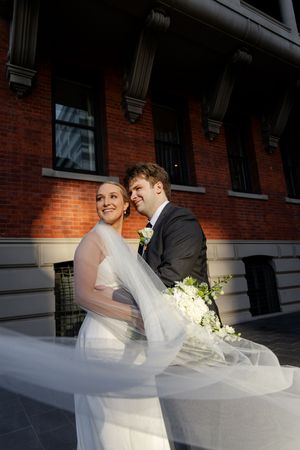 A bride and groom pose together outside a brick building as her veil flows in the breeze.