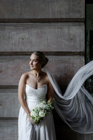 A flowing veil catches movement as someone poses in a strapless white wedding gown against an industrial concrete wall.