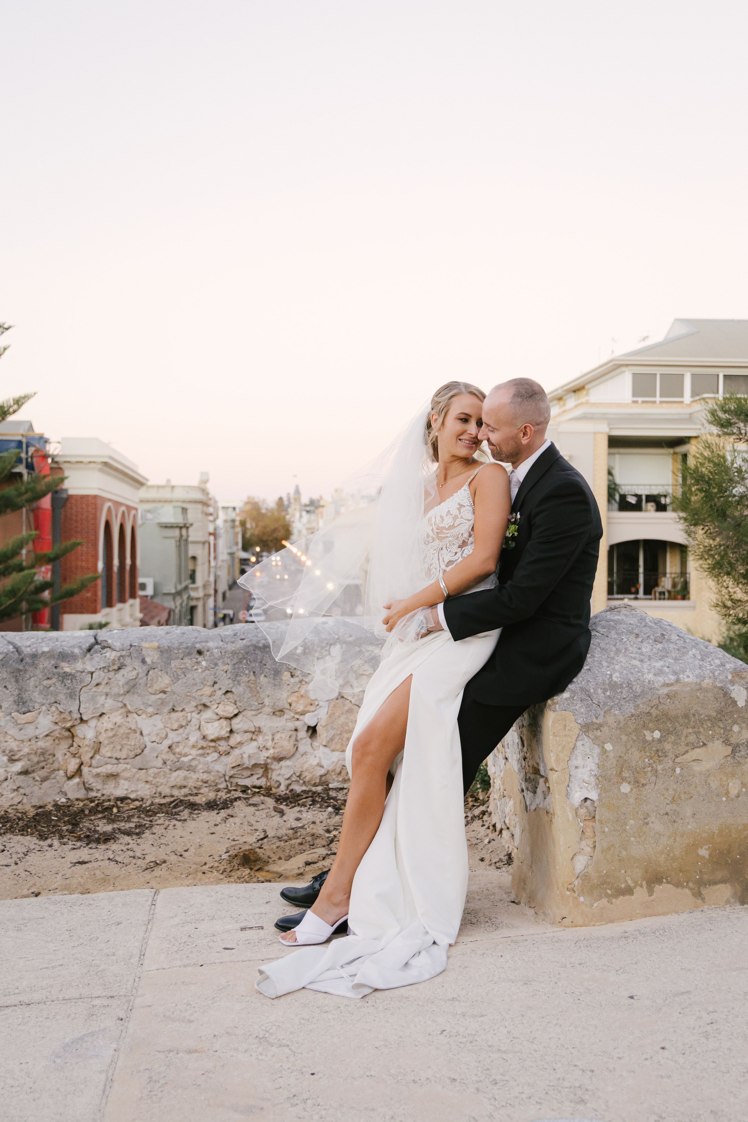 A romantic wedding couple embraces on a stone wall at sunset in an urban setting, bride wearing a flowing white gown.