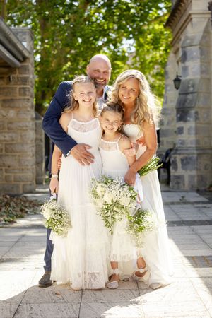 A joyful wedding party poses together on stone steps in bright sunlight with white dresses and floral bouquets.