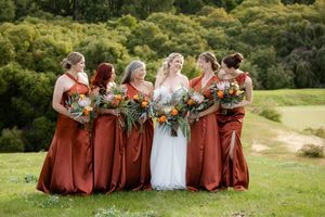 Wedding party wearing burgundy dresses poses together on grassy hillside with rustic floral bouquets.