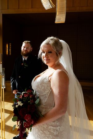 A bride in a white lace wedding dress holds a deep red bouquet while standing in warm sunlit room.