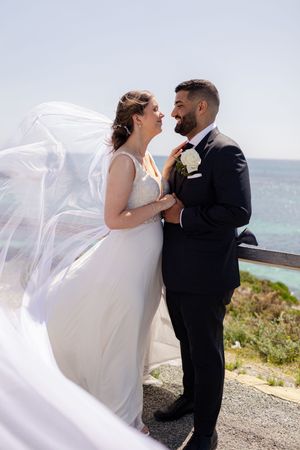 A romantic wedding couple shares intimate moments by the ocean as the bride's veil flows in the coastal breeze.