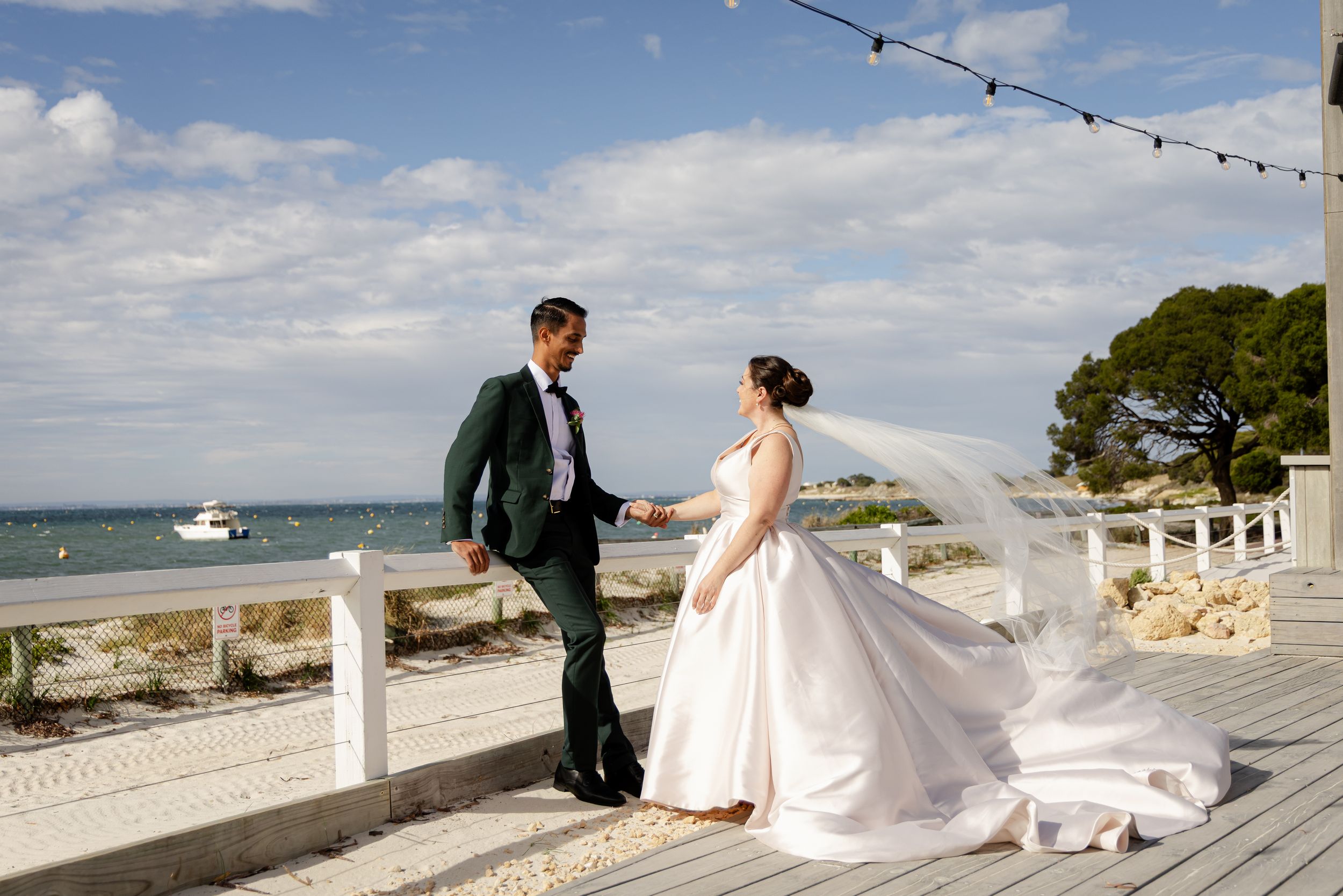 A couple in wedding attire stands on a seaside terrace with string lights overhead and boats in the distance.