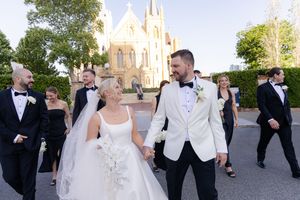 Wedding party in formal attire poses together outdoors with a church spire visible in the background.