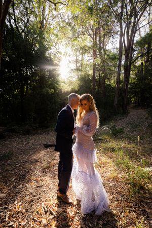 A couple in wedding attire walks through sunlit woods during golden hour as sunbeams filter through the trees.