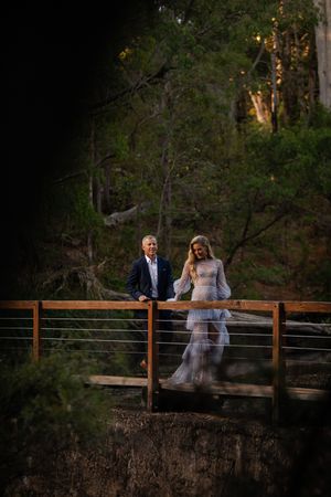 A couple sits together on a rustic wooden bridge surrounded by lush forest scenery at sunset.
