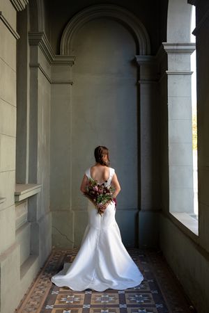 A bride in a white gown holds a bouquet while standing in an ornate columned corridor.