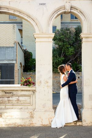 A romantic wedding couple embraces under elegant stone archways of a historic building.