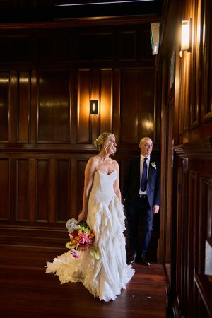 A bride and groom walk through an elegant wood-paneled hallway illuminated by warm wall sconces.