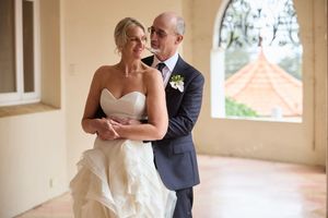 A couple in wedding attire dances together in a bright sunlit hallway with elegant columns and windows.