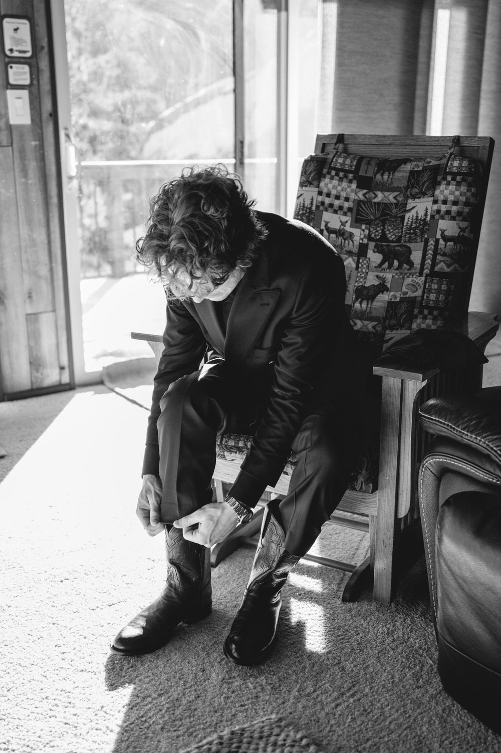 Black and white photo showing someone tying their shoes while sitting on a chair by a window.