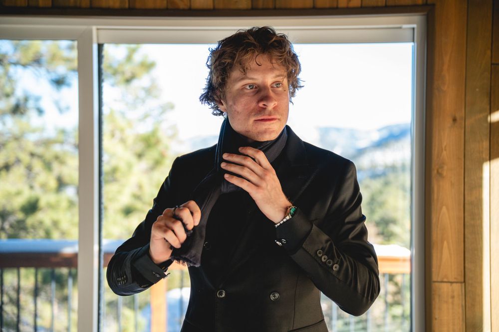 A person in a black coat adjusts their collar while standing by a window with mountain scenery in the background.